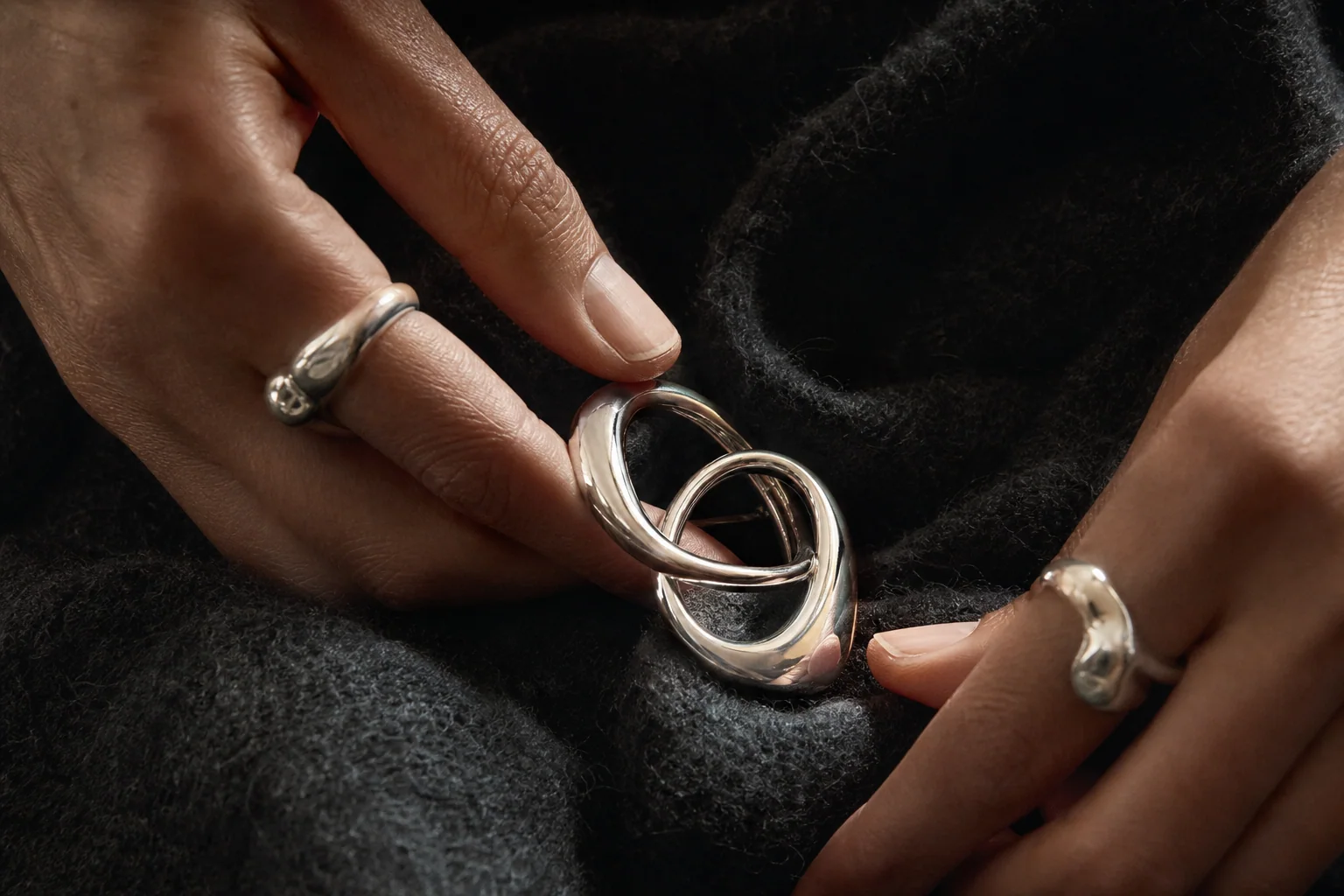 Hands styling sculptural silver jewelry over black wool